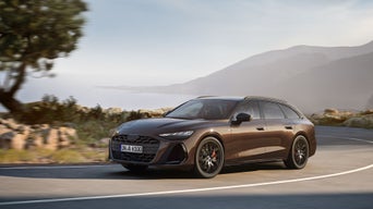 Ein brauner Audi fährt auf einer Küstenstraße, im Hintergrund Berge, Felsen und das Meer bei leichtem Dunst.