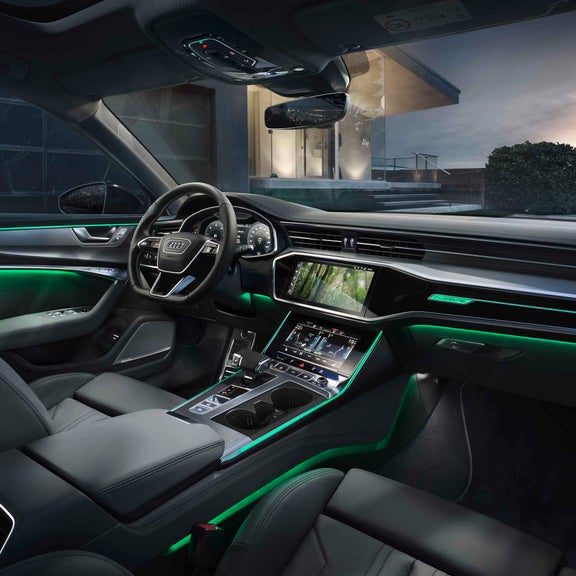 Audi Innenraum mit grünem Ambientelicht und digitalen Displays, im Hintergrund modernes Haus mit Treppe, Hecke und Sonnenuntergang.