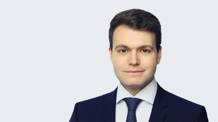 Markus Baumeister: Porträt jüngerer, freundlich schauender Mann mit dunklem Haar und Anzug vor grauem Hintergrund.