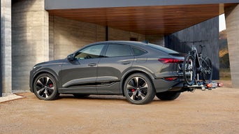 Grauer Audi Q6 e-tron mit zwei Fahrrädern auf Heckträger steht vor modernem Gebäude mit Holz- und Betonfassade.