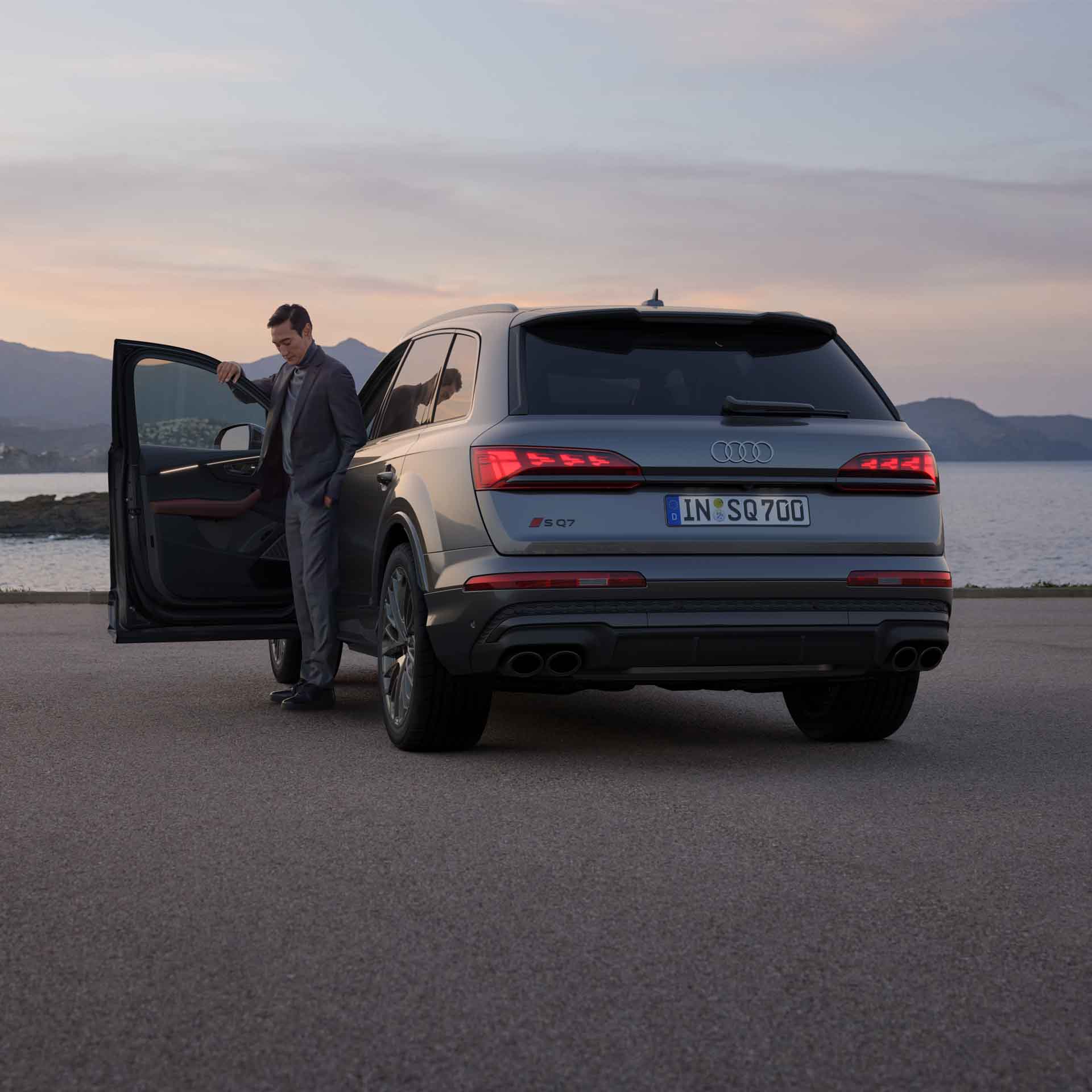 Ein grauer Audi SQ7 SUV (Rückansicht) steht an Uferstraße, Mann steigt bei Sonnenuntergang aus dem Fahrzeug, Berge im Hintergrund.