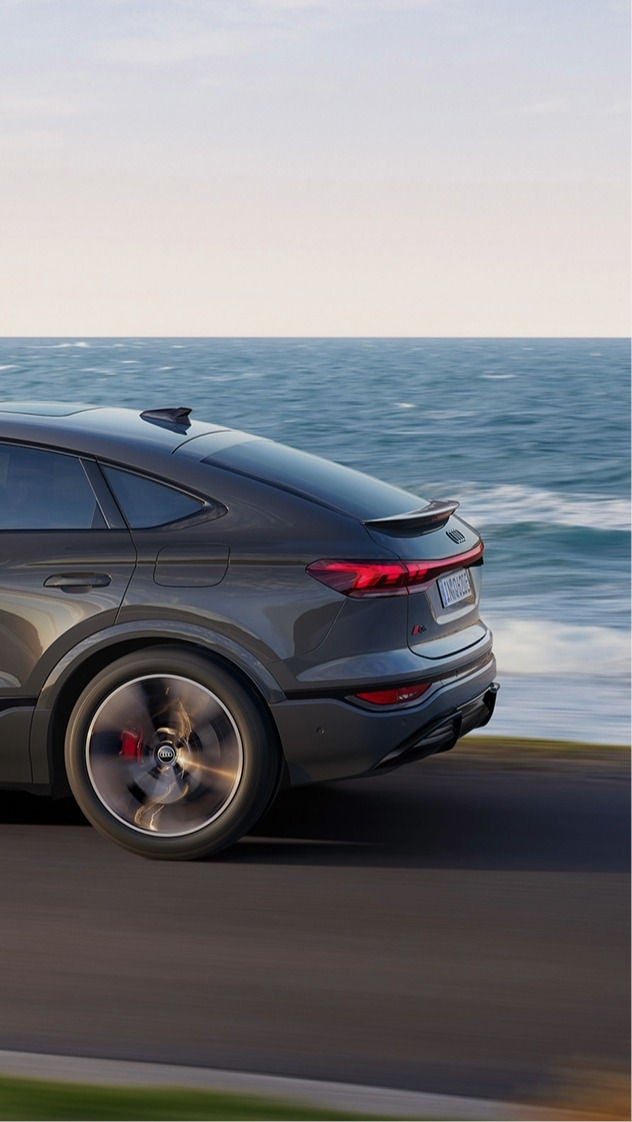 A grey Audi drives along a coastal road with ocean waves and cliffs in the background. The scene conveys a sense of freedom and adventure.