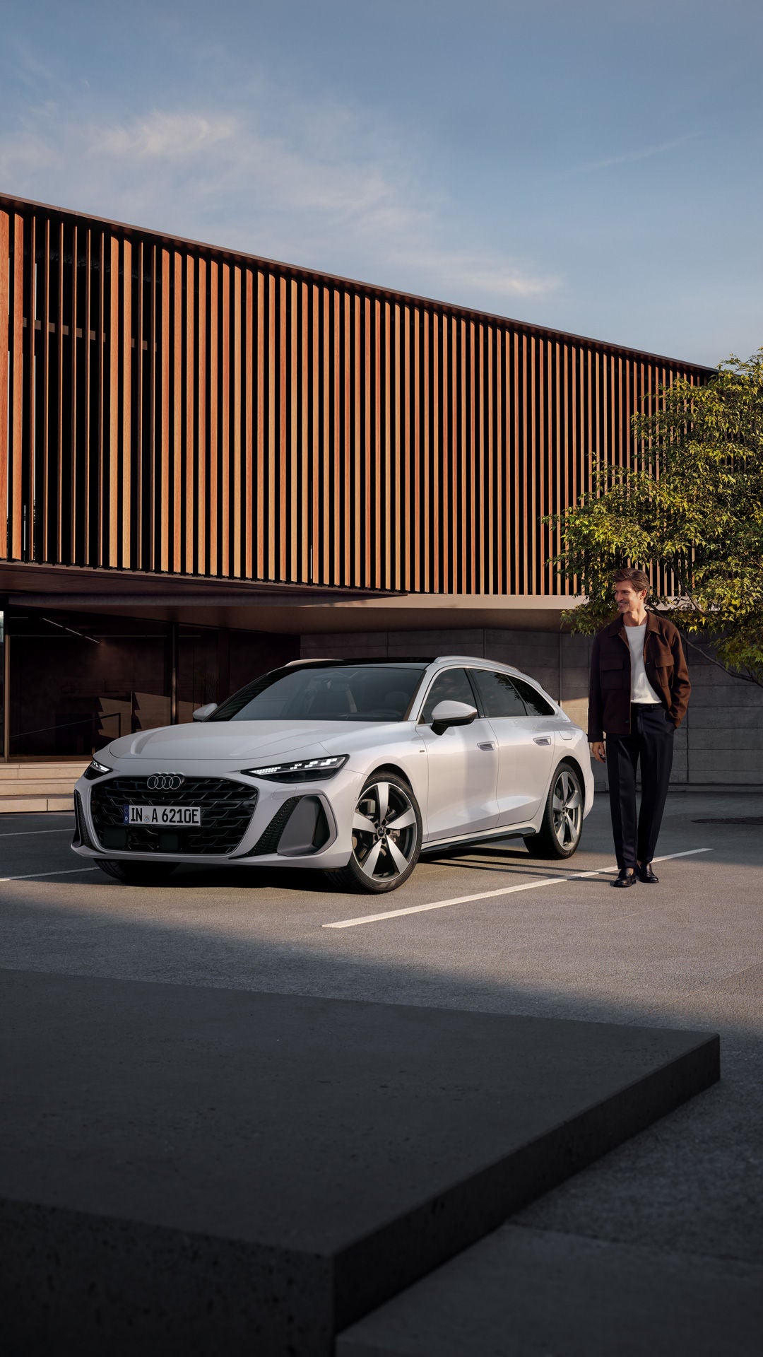Der A6 Avant e-hybrid steht im schrägen Frontprofil auf einem Parkplatz vor einem großen Gebäude mit Holzvertäfelung. Neben dem Fahrzeug steht ein Mann mittleren Alters.
