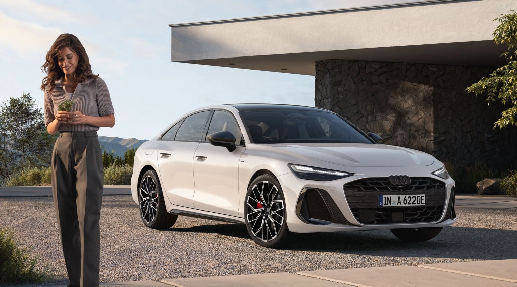 Die weiße A6 Limousine e-hybrid in schräger Frontansicht parkt vor einem modernen Steinhaus. Davor steht eine junge Frau, welche Blumen in der Hand hält.
