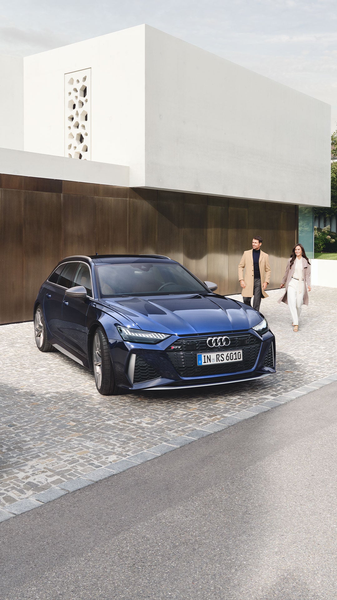 Dunkelblauer Audi RS 6 Avant parkt auf Kopfsteinpflaster, im Hintergrund ein Paar vor moderner Fassade.