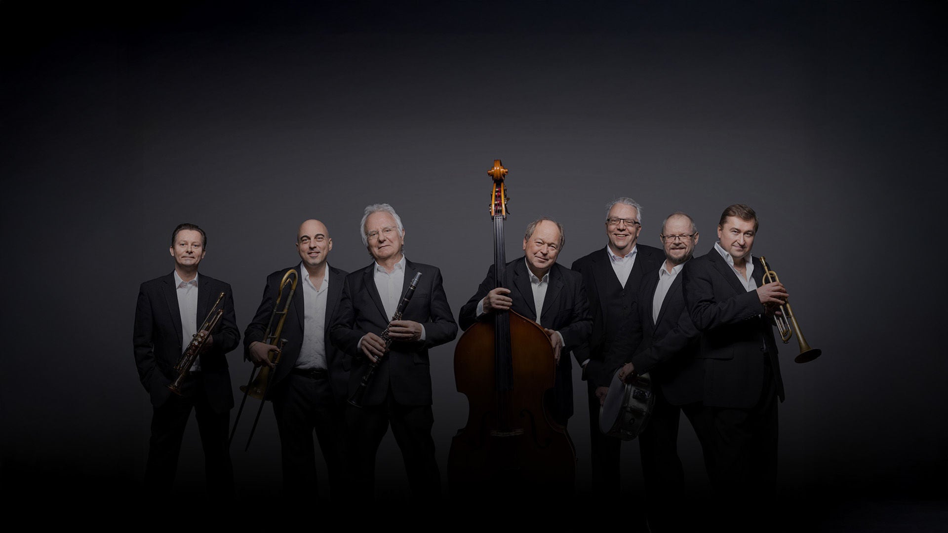 Sieben Jazzmusiker in dunklen Anzügen posieren vor grauem Hintergrund mit Trompeten, Posaune, Klarinette, Kontrabass und Schlagzeug.