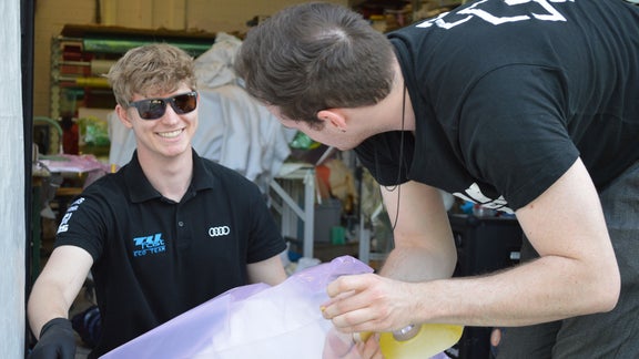 A man has his hands on a piece in a plastic bag and looks at another man who is smiling at him and wearing sunglasses