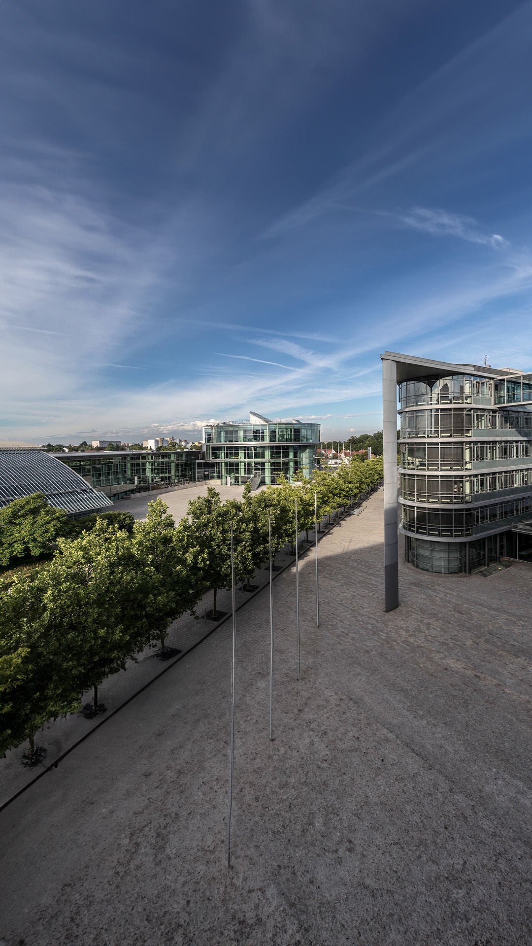 Blick auf moderne Gebäude mit Glasfassaden, breitem Vorplatz und begrüntem Bereich bei Tageslicht.