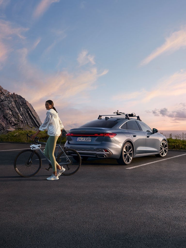 Eine Frau steht mit einem Fahrrad neben einem geparkten silbernen Audi A5 Limousine vor einer Felsformation bei Sonnenuntergang.