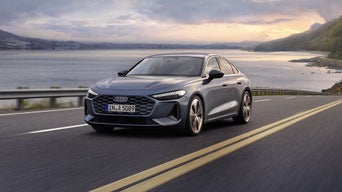 Sleek grey car on a scenic coastal road during sunset. The sea and hills are in the background.