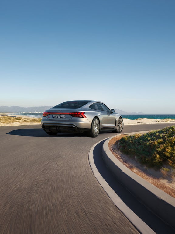 Ein silberner Audi S e-tron GT fährt auf einer kurvigen Küstenstraße mit Blick auf das Meer und eine Stadt im Hintergrund.