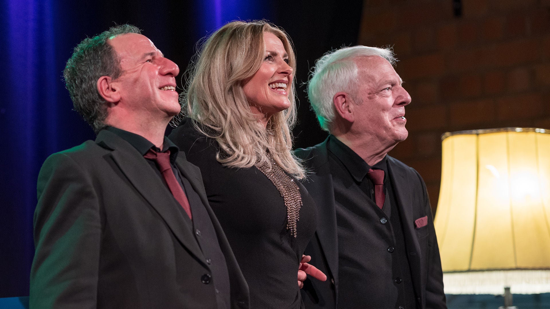 Drei Musiker in schwarzen Anzügen lächeln auf einer Bühne unter farbigem Bühnenlicht neben einer Stehlampe.