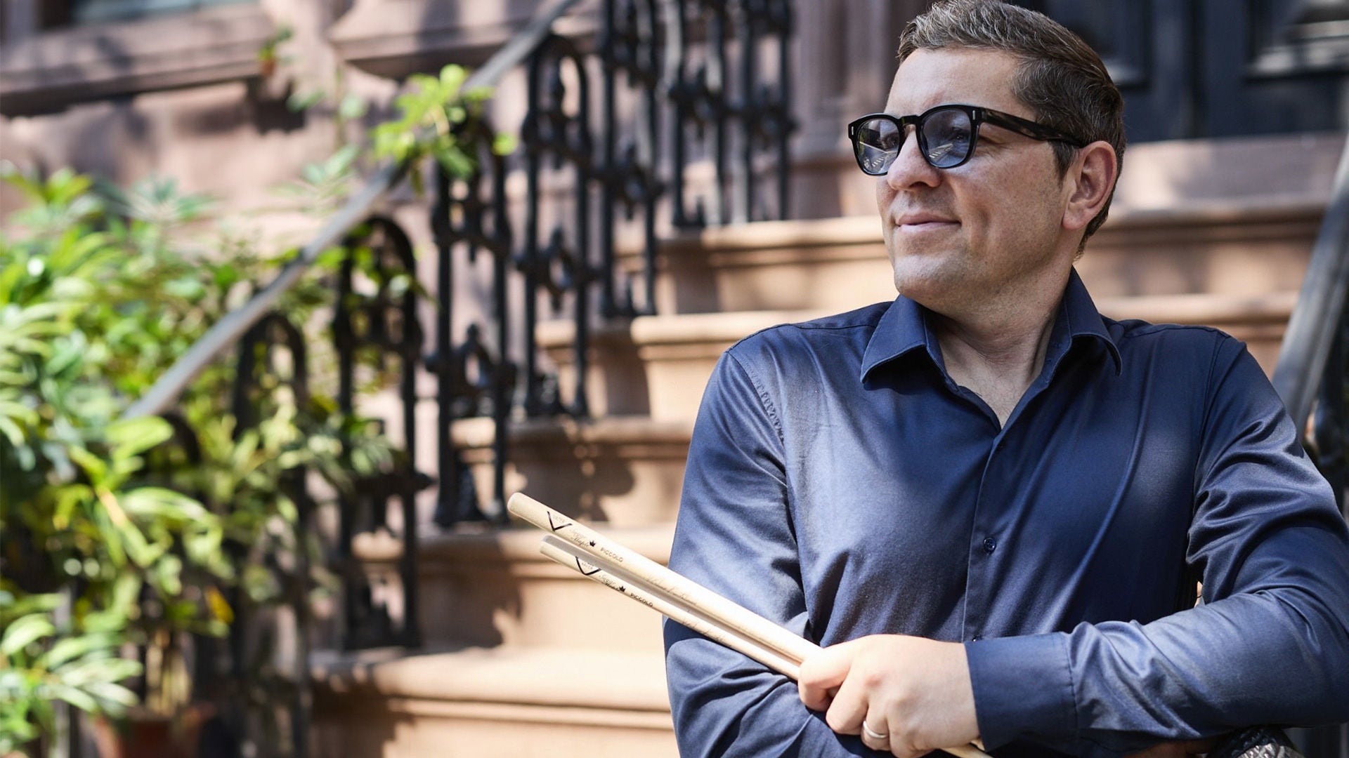 Schlagzeuger von Iris Quintett steht auf einer Treppe an ein Geländer gelehnt mit Drums Sticks in der Hand. Trägt eine Sonnenbrille das Wetter ist frühlingshaft. Die Sonne scheint.