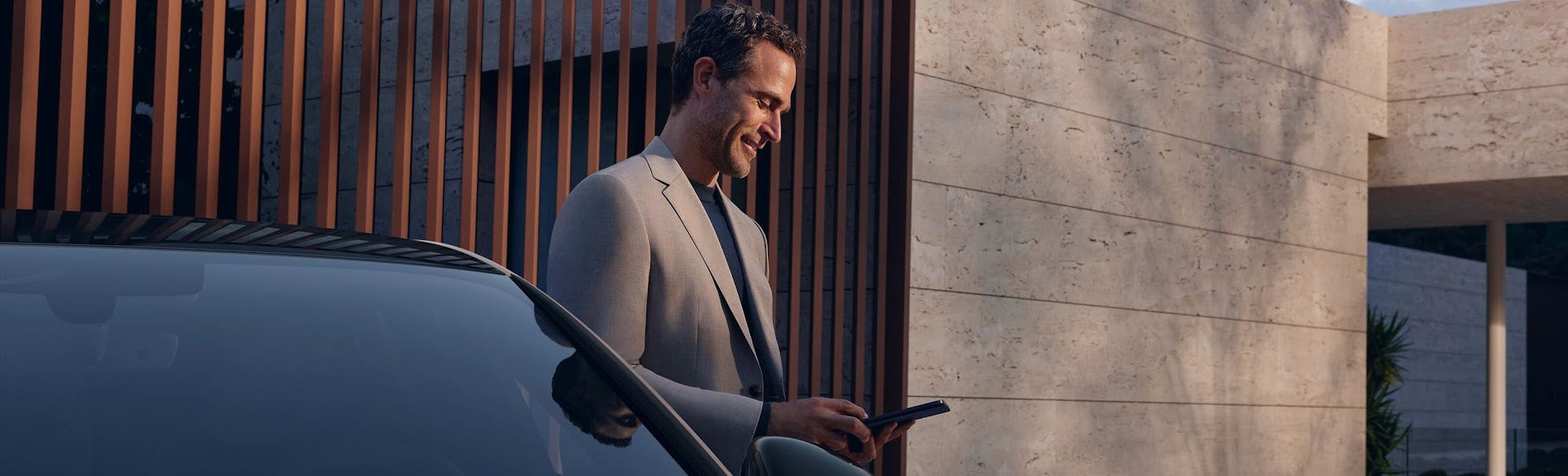 A man stands by a car, checking his phone with a modern architectural backdrop of stone and wooden slats.