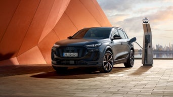 Seitliche Front des SQ6 SUV e-tron, welcher neben einer orangenen Gebäudefassade an eine Ladestation angeschlossen ist. Im Hintergrund ist das Meer, sowie die Skyline einer Stadt zu erkennen.