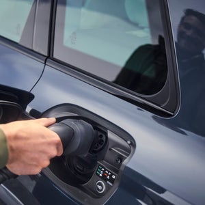 A person plugs a charging cable into a sleek electric car, reflecting sustainability. The glossy car exterior shows a faint reflection of the individual.