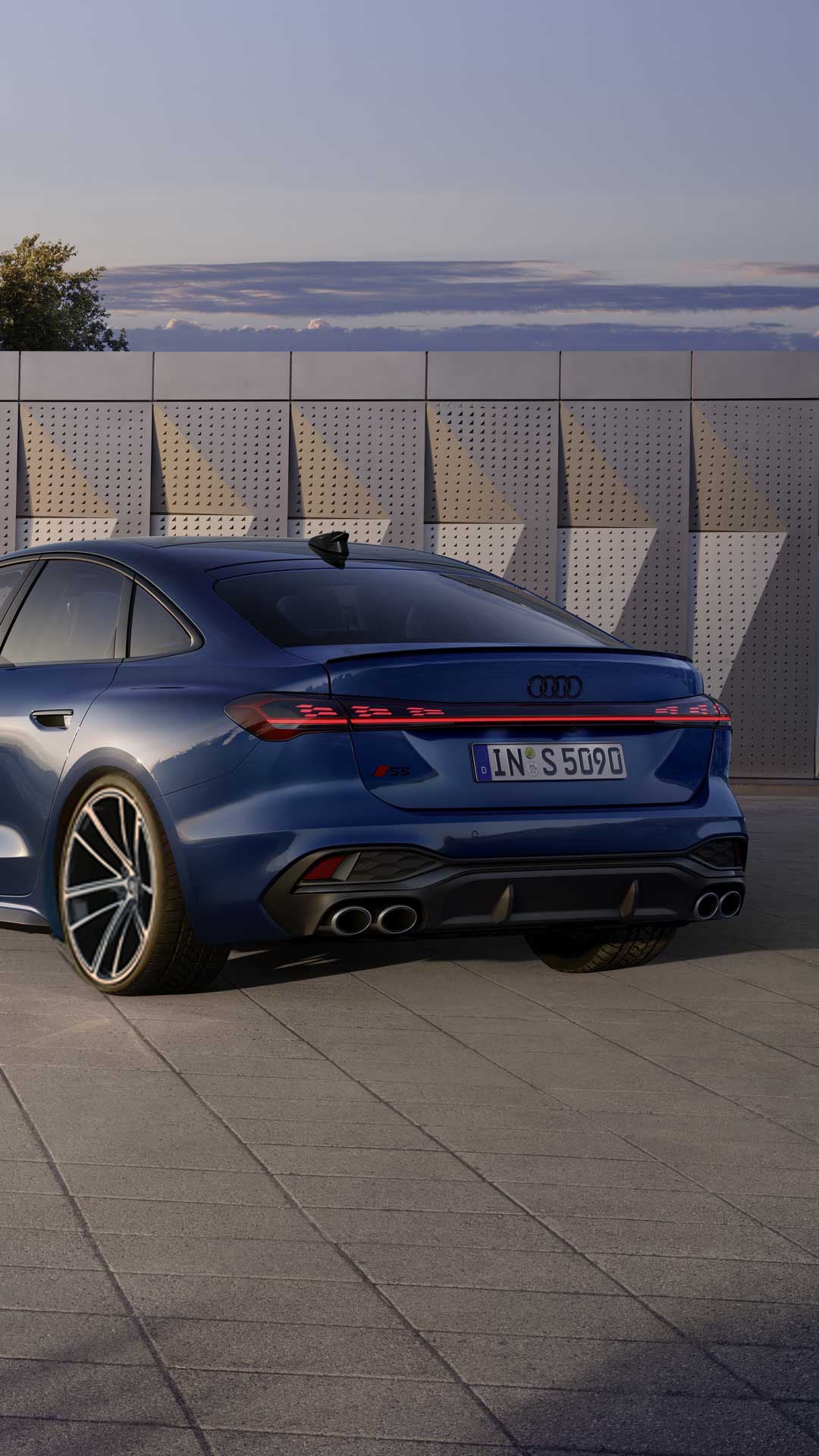 The dark blue S5 Saloon is parked at an angle in front of a building facade and is illuminated by sunlight.