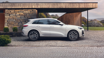 A white SUV parked beside a modern stone and wood house, set against a scenic backdrop of hills and clouds.