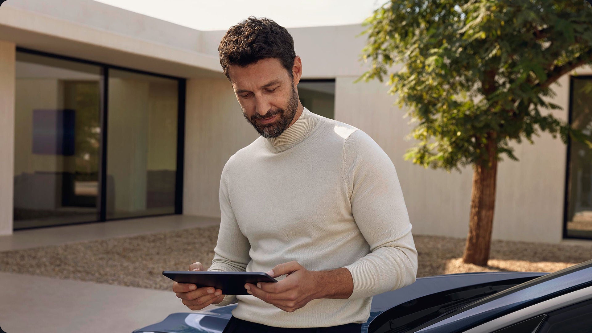 A man with a beard, in a light sweater, looks at a tablet while leaning on a car. He's outdoors near a modern building and a tree, appearing focused.