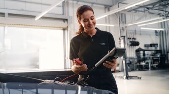 Mechanikerin in schwarzer Audi-Uniform prüft Fahrzeug mit Diagnosegerät in heller Werkstatt.
