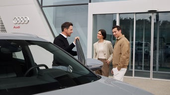 Mann zeigt zwei Personen ein Auto vor Audi Gebäude, sie stehen neben grauem Fahrzeug vor Glasfassade.