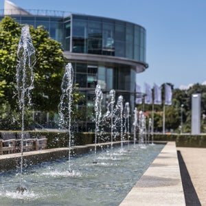 Wasserfontänen in langem Becken, im Hintergrund rundes Glasgebäude und grüne Bäume bei blauem Himmel.