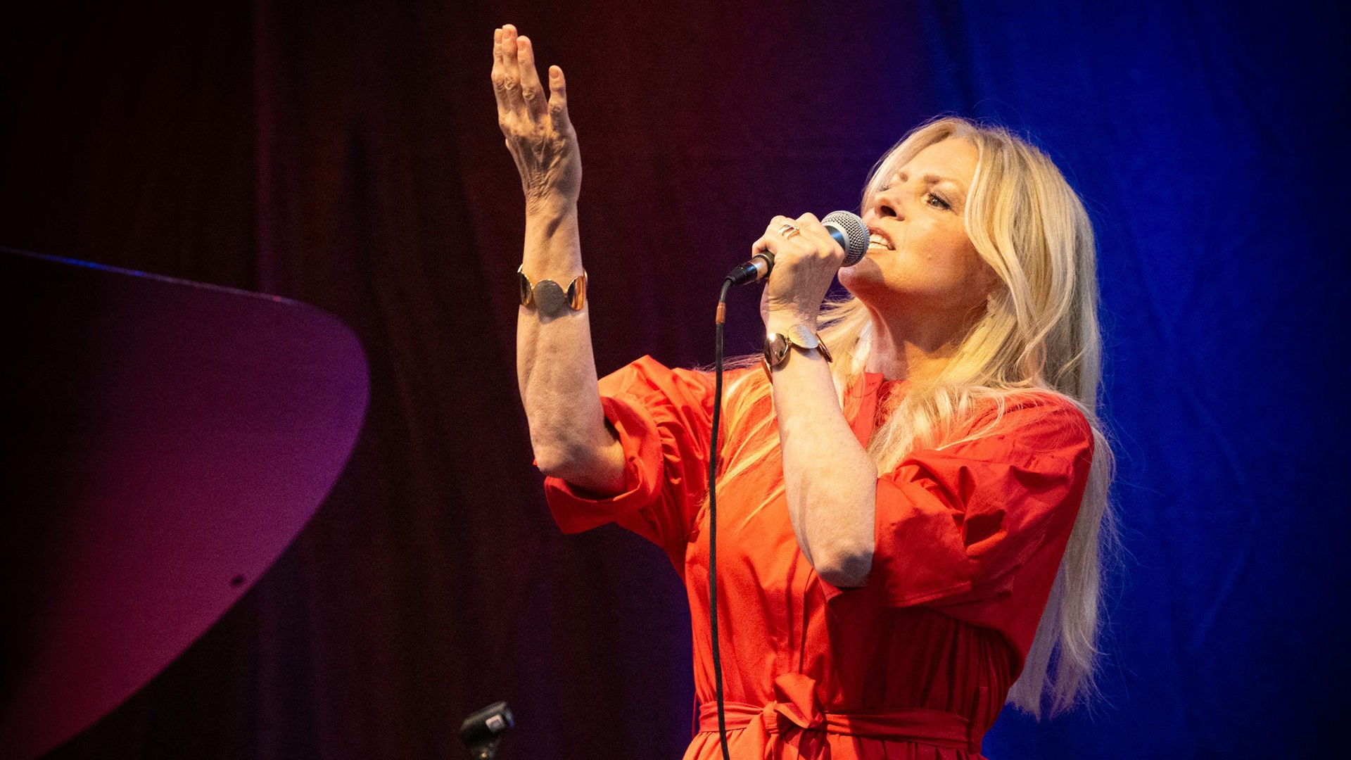 Blonde Frau in rotem Kleid singt ins Mikrofon und hebt einen Arm vor blau-violettem Bühnenlicht.