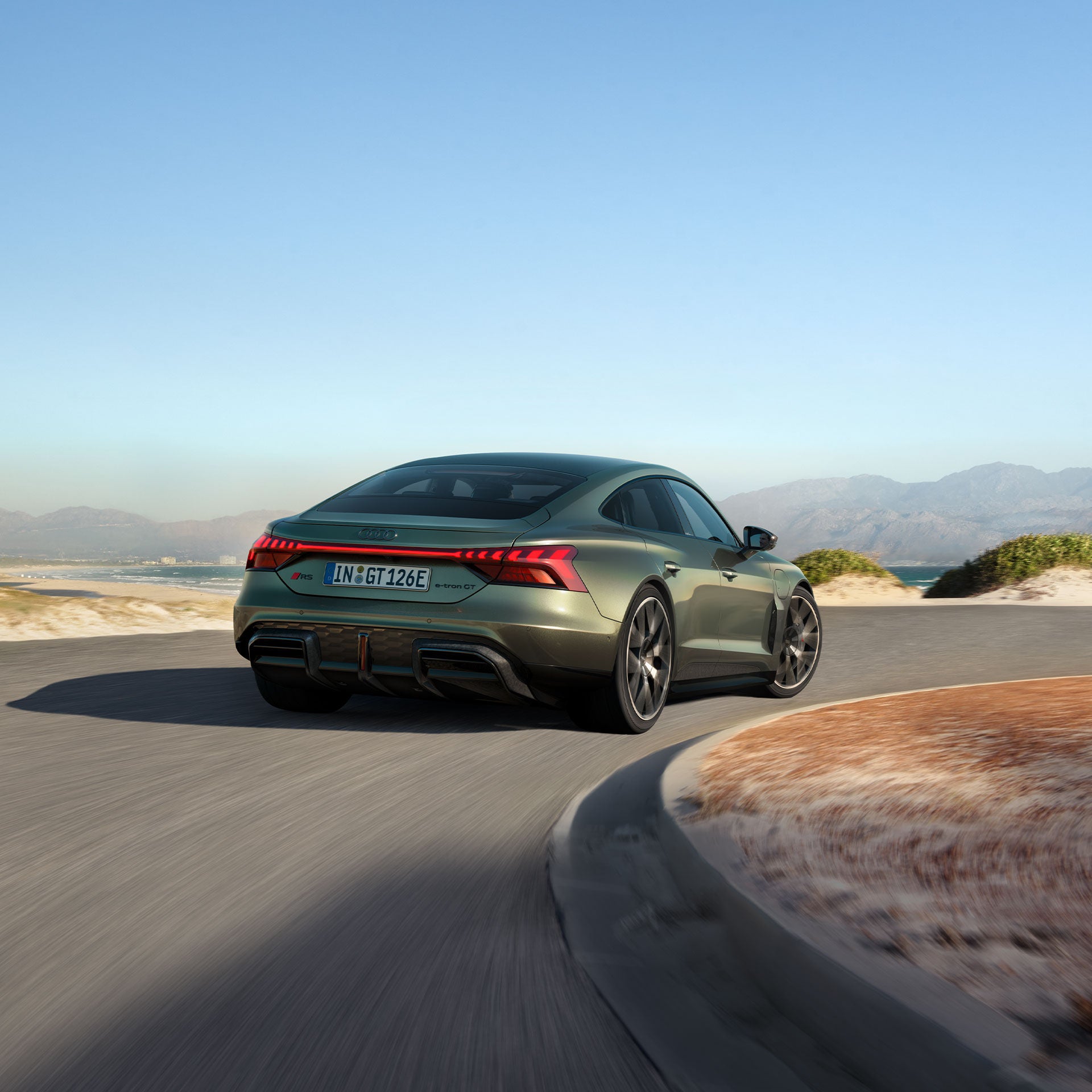 Grüner Audi RS e-tron GT performance fährt auf einer kurvigen Küstenstraße mit Meer, Dünen und Bergen im Hintergrund.