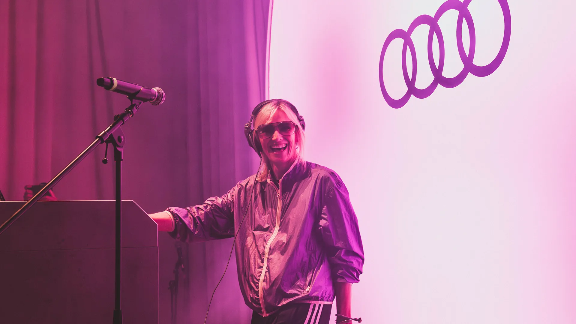 A DJ in sunglasses and headphones smiles at her decks, bathed in pink and purple stage lighting beside the glowing Audi logo. A DJ in sunglasses and headphones smiles at her decks, bathed in pink and purple stage lighting beside the glowing Audi logo.
