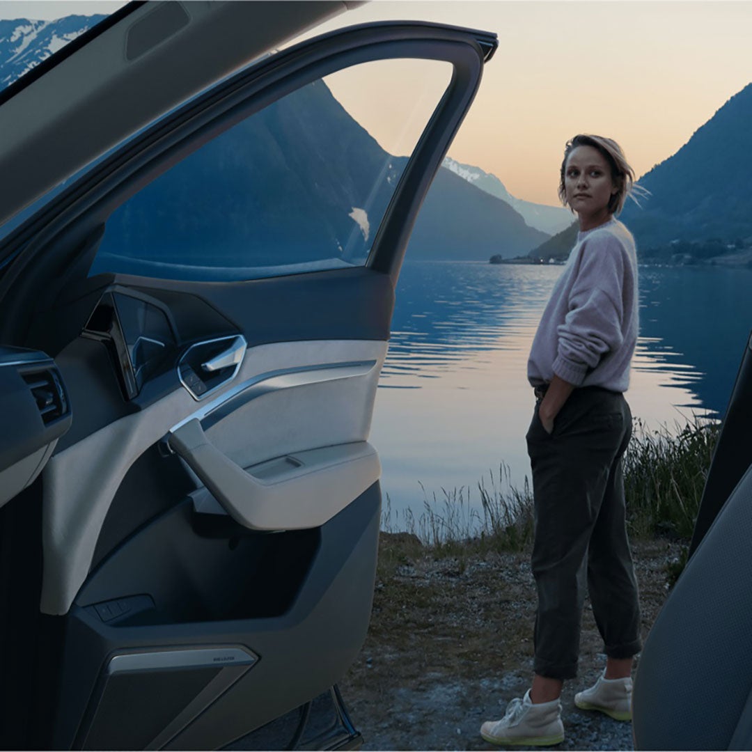 Frau steht am See neben offener Autotür; Berge im Abendlicht, ruhiges Wasser.