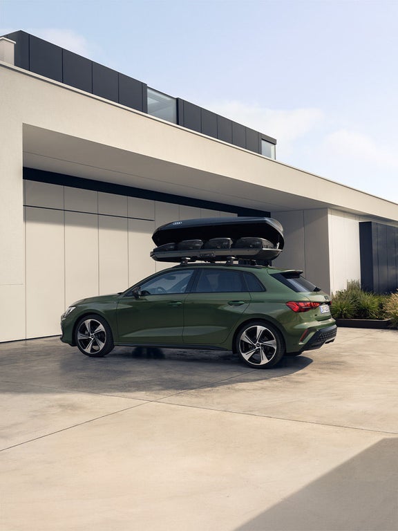 Grüner Audi A3 Sportback mit offener Dachbox, geparkt vor einem modernen weißen Gebäude mit Pflanzen im Hintergrund.