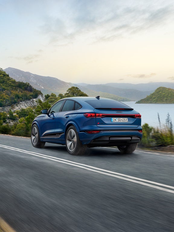 Heckansicht eines blauen Audi SQ6 Sportback e-tron auf Landstraße, im Hintergrund grüne Hügel und ein großer See bei Tageslicht.