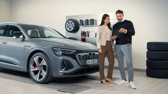 Zwei Personen stehen vor einem grauen Audi e-tron in einer Werkstatt und betrachten gemeinsam ein Tablet, Reifen im Hintergrund.