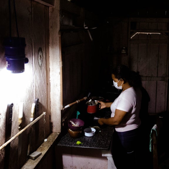 Eine Frau steht am Herd und bereitet Essen zu. Der dunkle Raum wird durch eine Solarlichtquelle erhellt.