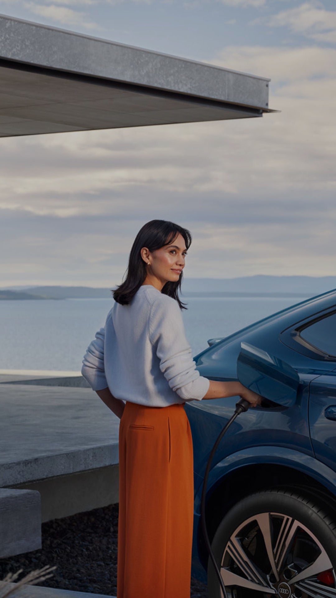 A woman plugs an electric vehicle into a charger outside a modern home with a scenic view of the water and mountains in the background.