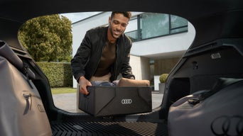 Mann stellt Audi Kofferraumbox mit Kleidung in geöffneten Kofferraum vor Haus.