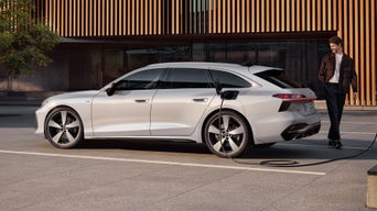 Der A6 Avant e-hybrid steht seitlich vor dem Eingangsbereich eines großen Gebäudes und wird über ein Stromkabel geladen. Am Heck des Fahrzeugs steht ein Mann mittleren Alters.