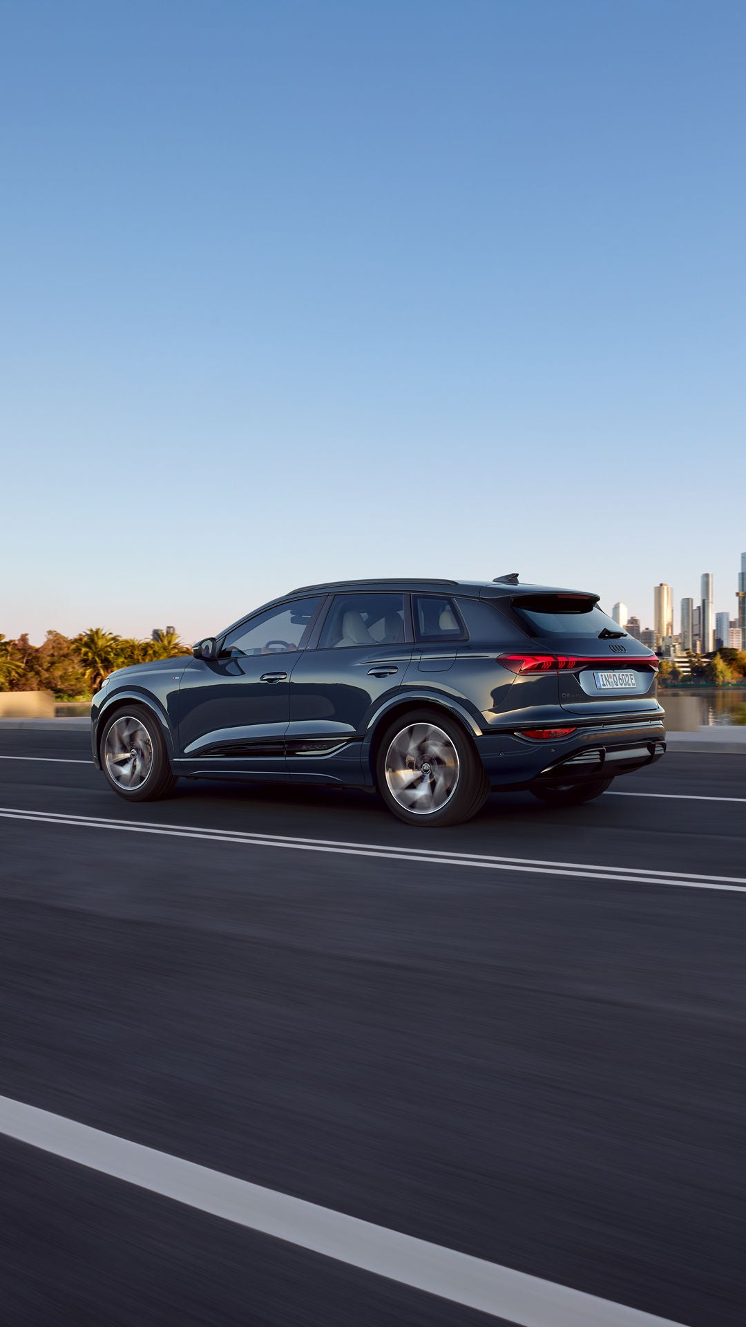 Zijprofiel van de Q6 e-tron rijdend op een weg langs een rivier.  Op de achtergrond is de skyline van een stad te zien.