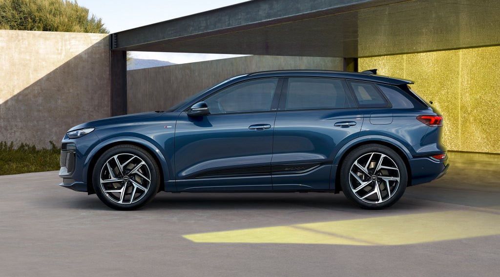 Der Q6 SUV e-tron in dunkelblau steht im Seitenprofil vor einem urbanen Haus. Im Vordergrund, leicht neben dem Auto steht eine junge, modische Frau.