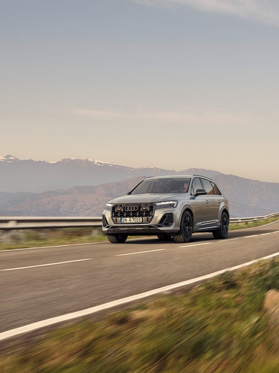 Ein silberner Audi Q7 SUV TFSI e fährt auf einer kurvigen Bergstraße bei Tageslicht, Berge im Hintergrund.