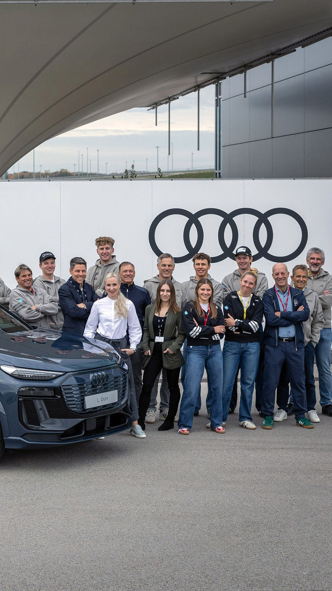 DSV-Top-Athletinnen und -Athleten am Audi Forum Ingolstadt vor einem grauen Audi Modell im Hintergrund sind rechts obern auf einer Wand die schwarzen Audi Ringe zu sehen.