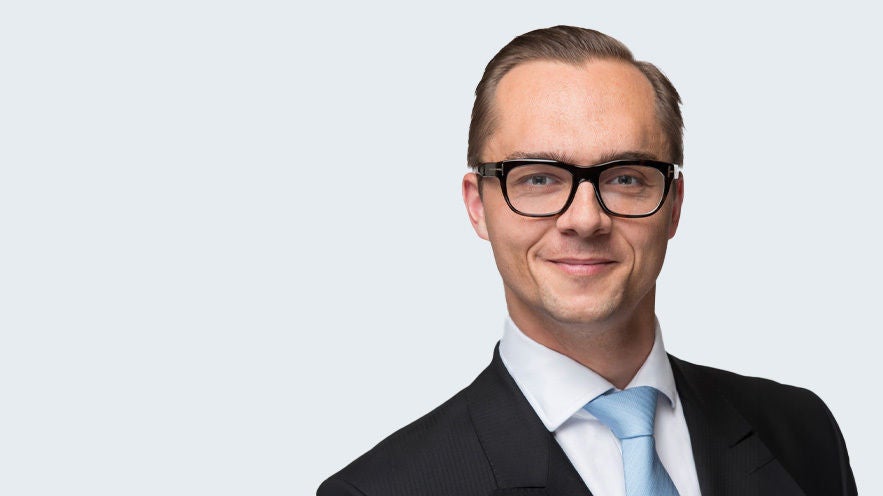 Dr. Marc Ruttloff: Porträt junger lächelnder Mann mit Brille, Kurzhaarschnitt und Anzug vor grauem Hintergrund.