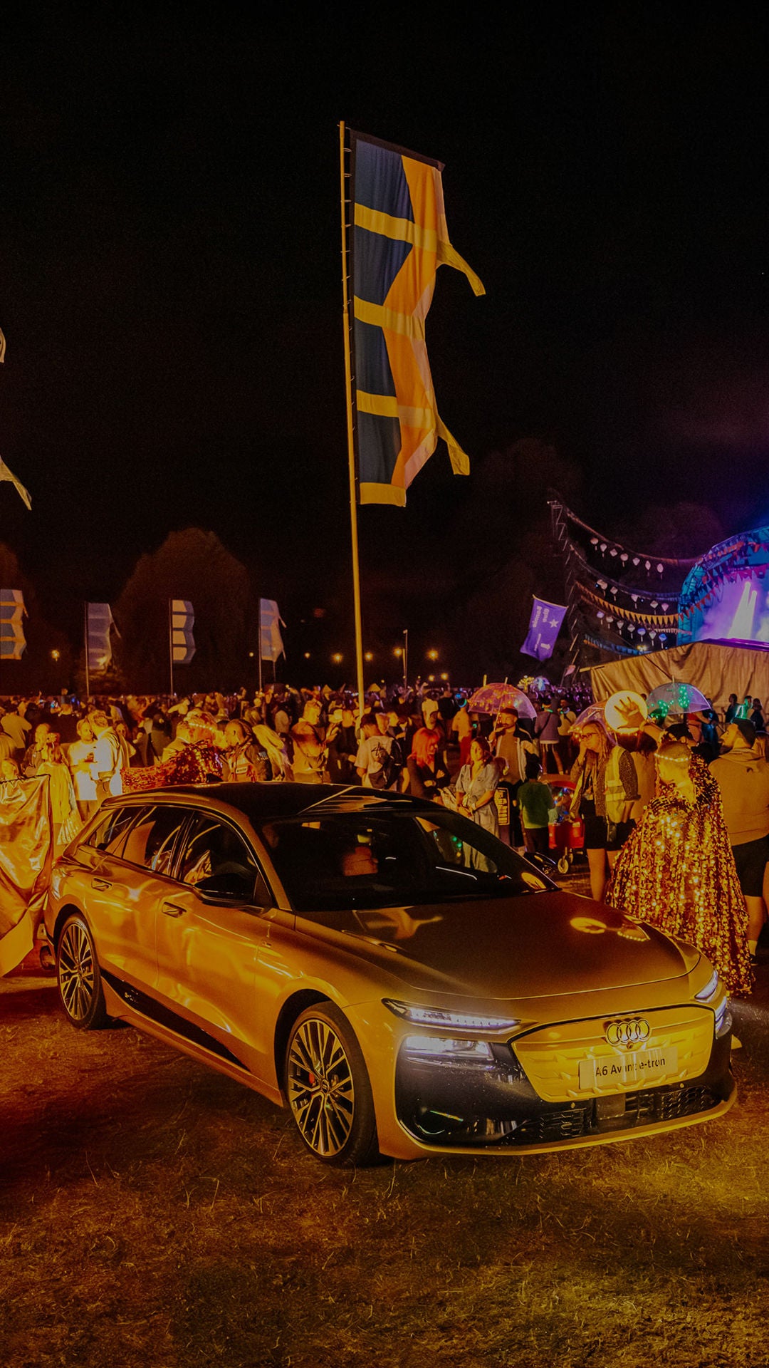 An Audi SQ6 e-tron at an outdoor event. Large winged structures and colorful flags adorn the background.
