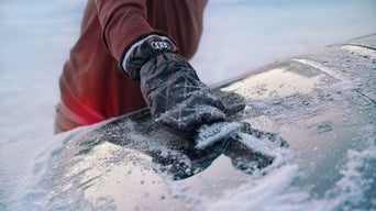 Hand mit Handschuh kratzt Eis von einer gefrorenen Autoscheibe.