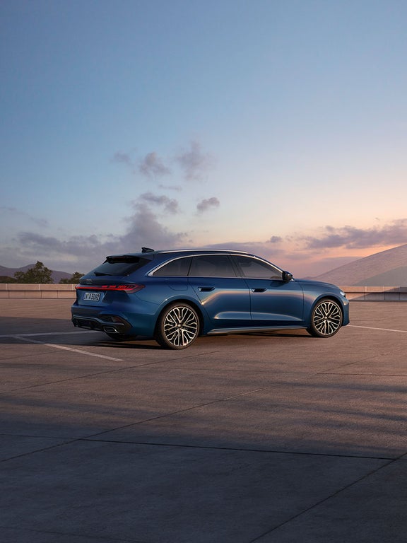 Eine Frau steht neben einem blauen Audi A5 Avant auf einem offenen Platz bei Sonnenuntergang.