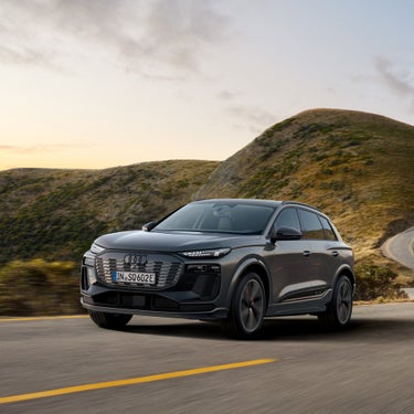 Frontansicht eines fahrenden, grauen Audi SQ6 SUV e-tron auf kurviger Bergstraße bei Sonnenuntergang.