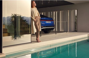 A modern outdoor space features a sleek blue Audi car behind a woman in stylish attire, beside a sparkling pool and a reflective glass wall.