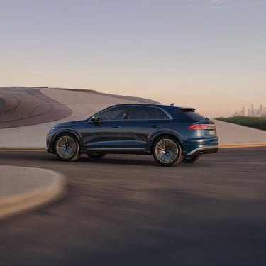 Ein dunkelblauer Audi SQ8 SUV fährt auf moderner Straße mit geschwungener Architektur im Hintergrund.