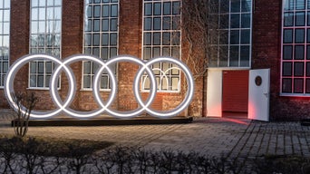 Im Hintergrund befindet sich ein altes Fabrik-Backsteingebäude mit hohen weissen Sprossenfenstern. Aus der geöffneten Tür dringt ein pinkfabener Lichtstrahl hervor. Im Vordergrungd befindet sich lebensgroße weiss leuchtende Audi Ringe.