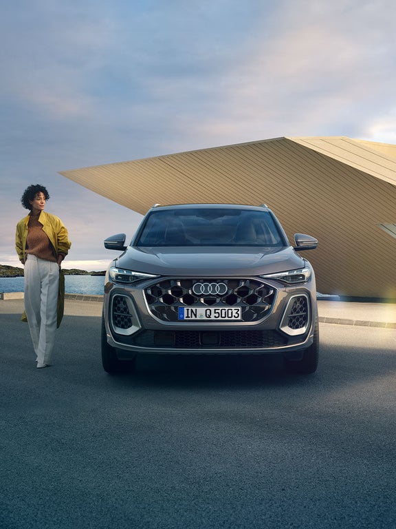 Ein grauer Audi Q5 SUV von vorne, daneben steht eine farbige Frau mit Afrohaar in gelber Jacke und heller Hose, im Hintergrund eine Küstenlandschaft.